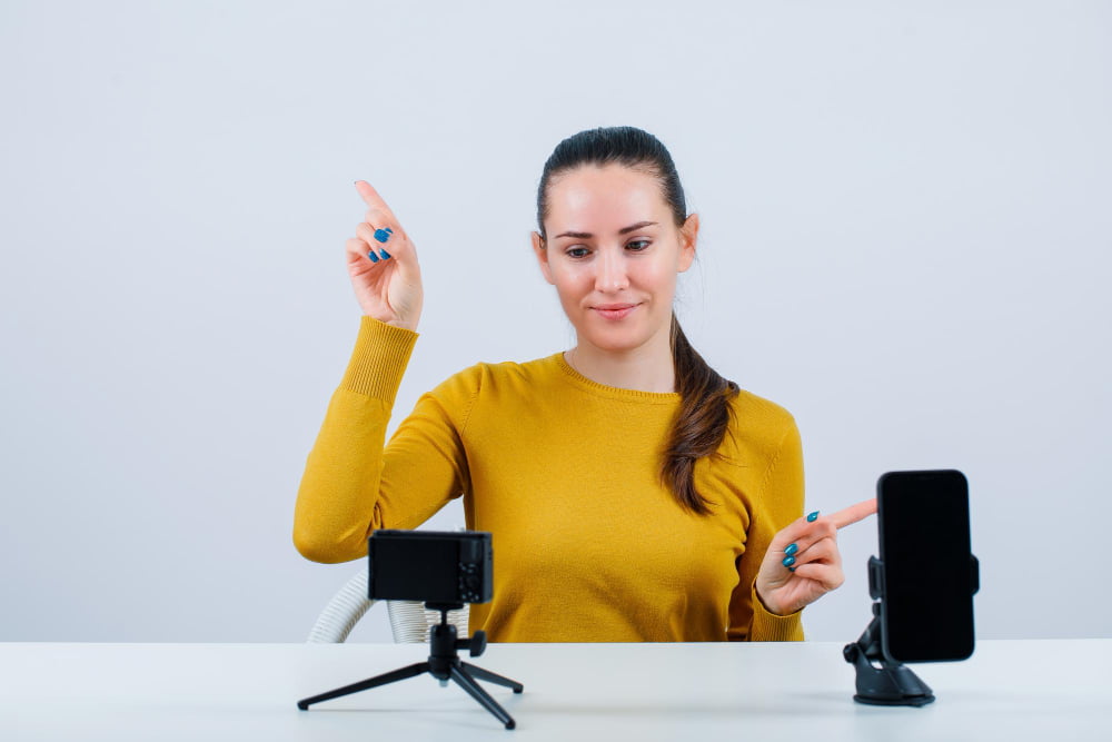 Como usar estratégia de vídeo para e-commerce? 3 smiling blogger girl is pointing up right with forefingers by sitting front her mini camera white background